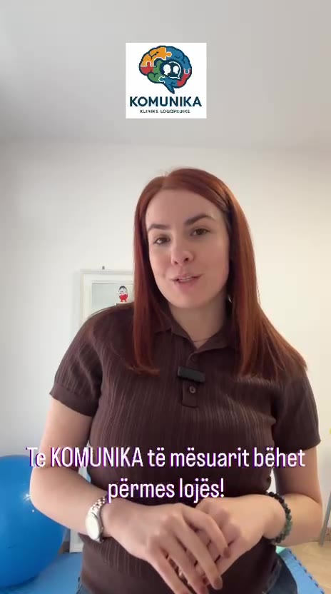 Një grua flet drejt kamerës, nën logon e KOMUNIKA; në fund të kuadrit shihet titra “Te KOMUNIKA të mësuarit bëhet përmes lojës!”